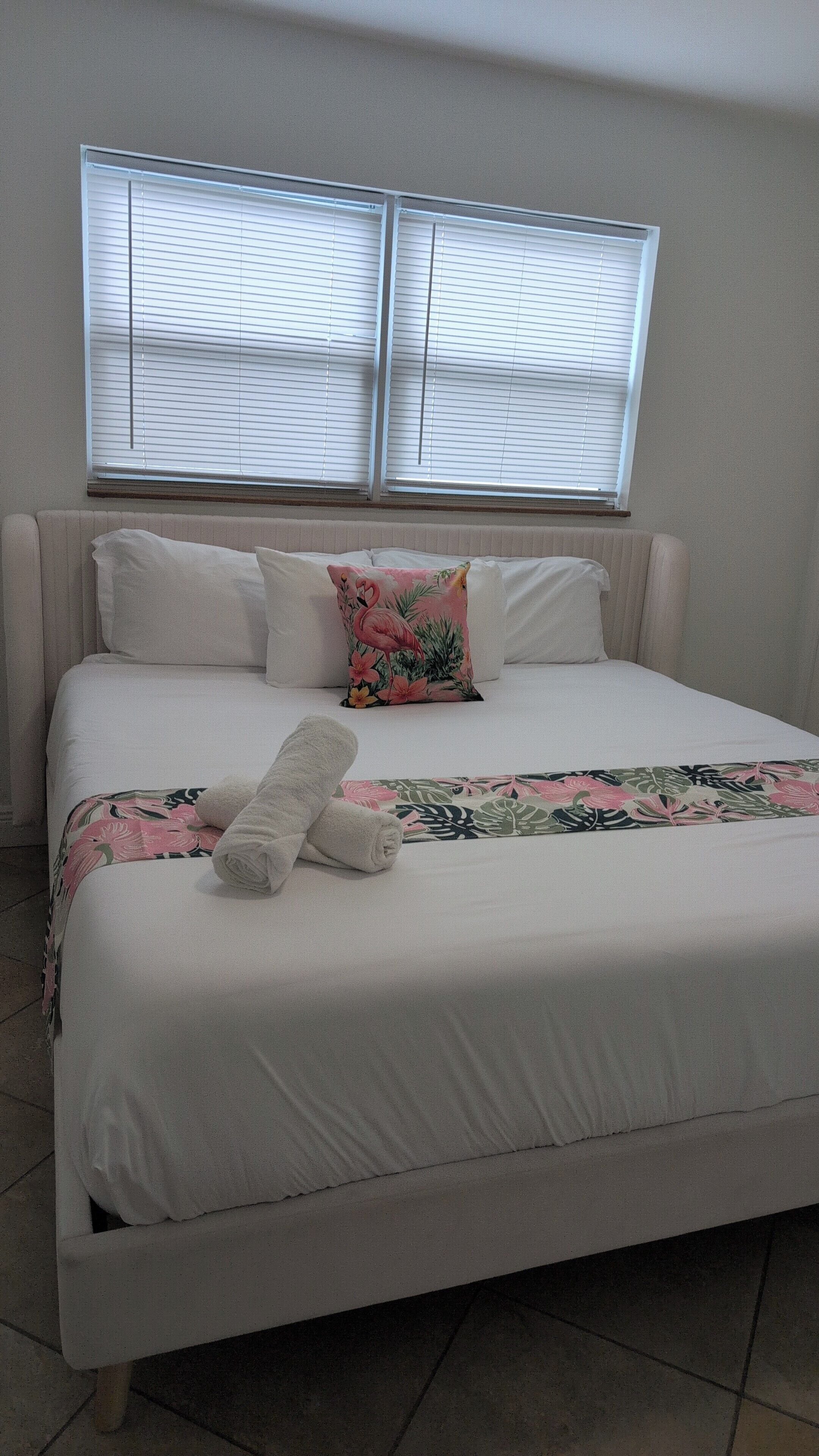 Una cama con ropa de cama blanca, un cojín floral y una toalla enrollada.