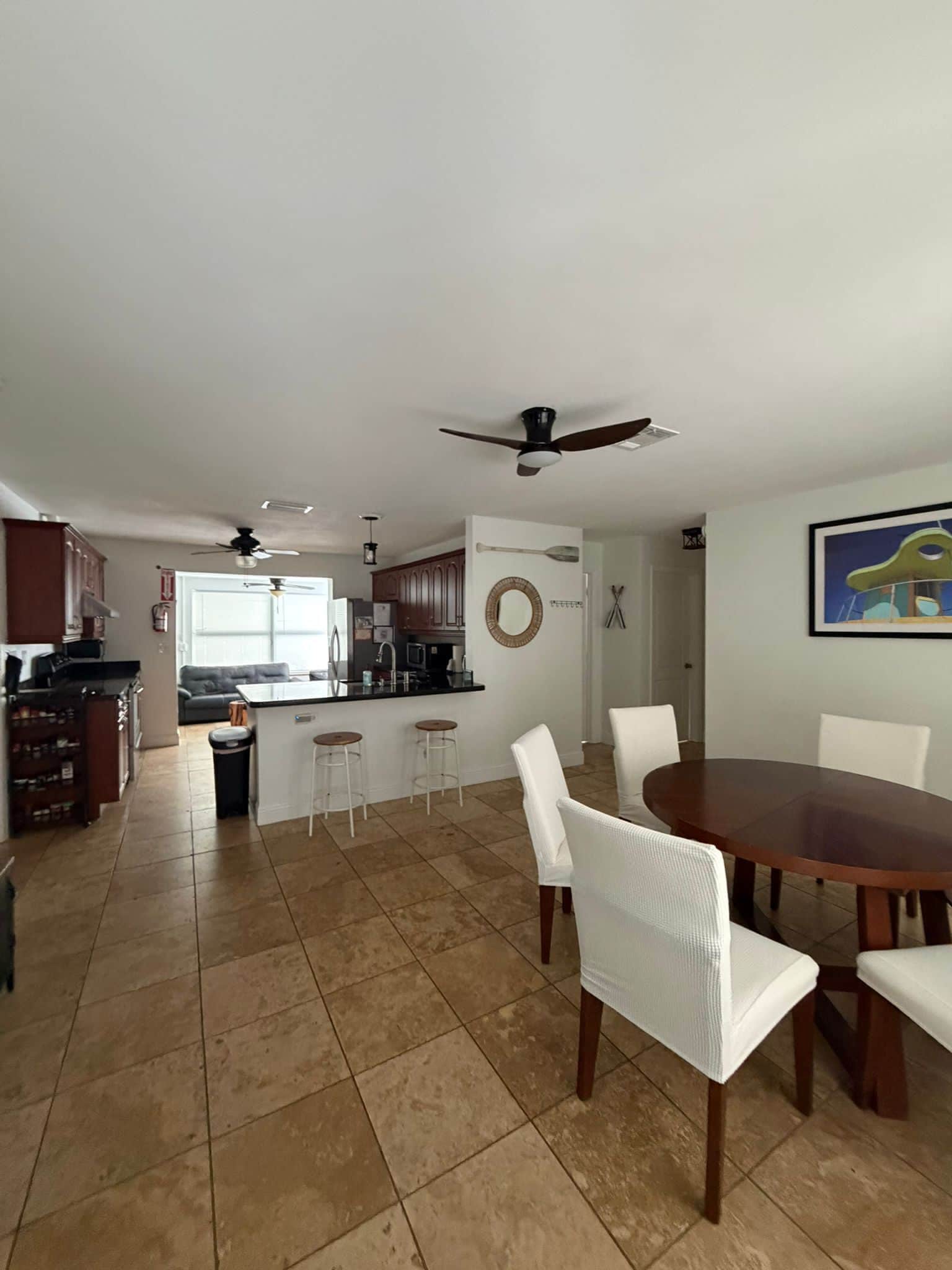 A spacious living area with a dining table, white chairs, a kitchen with a bar, and a ceiling fan.