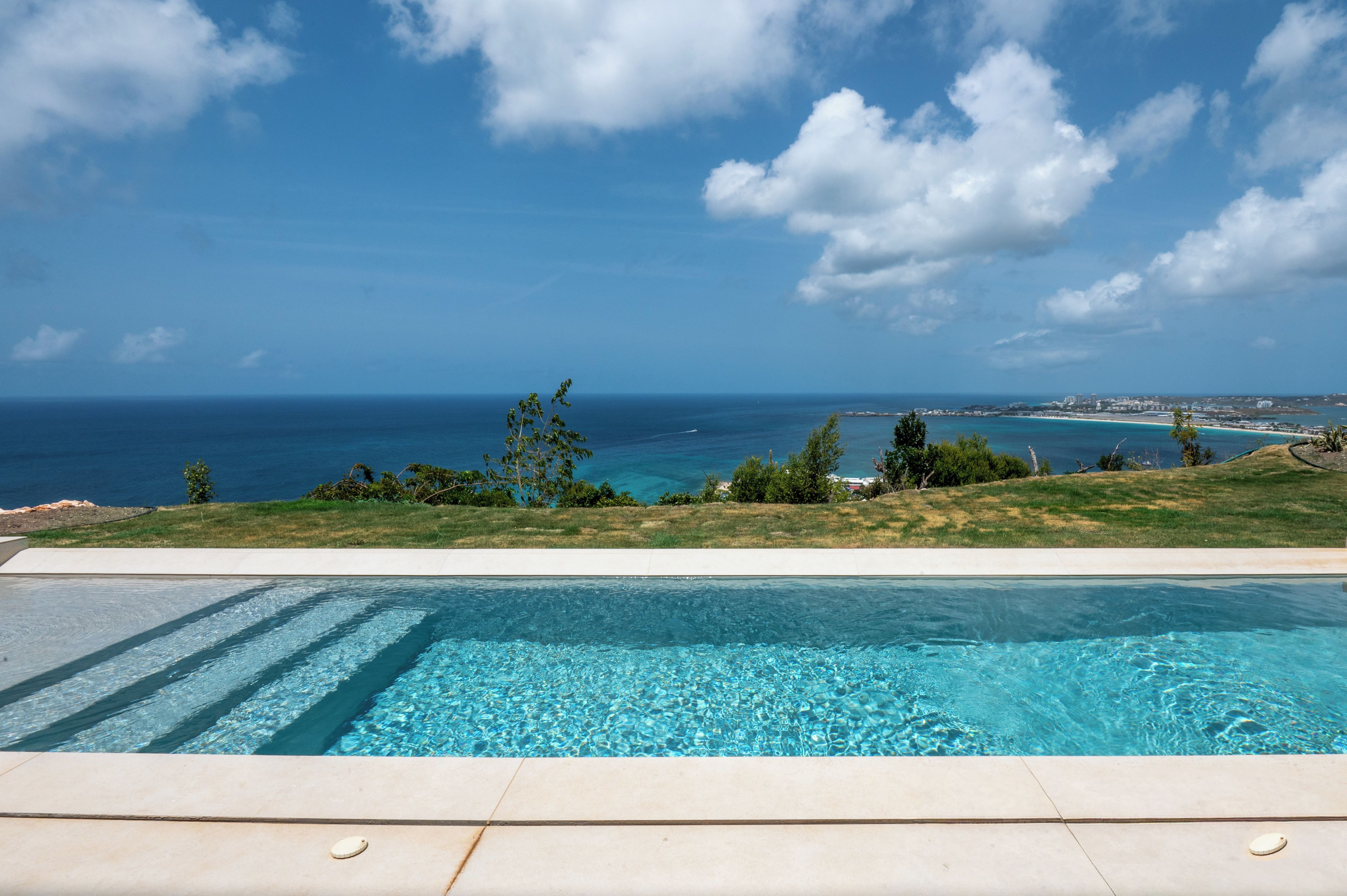 A swimming pool with clear blue water, surrounded by a grassy area with trees and a view of the ocean and distant buildings.