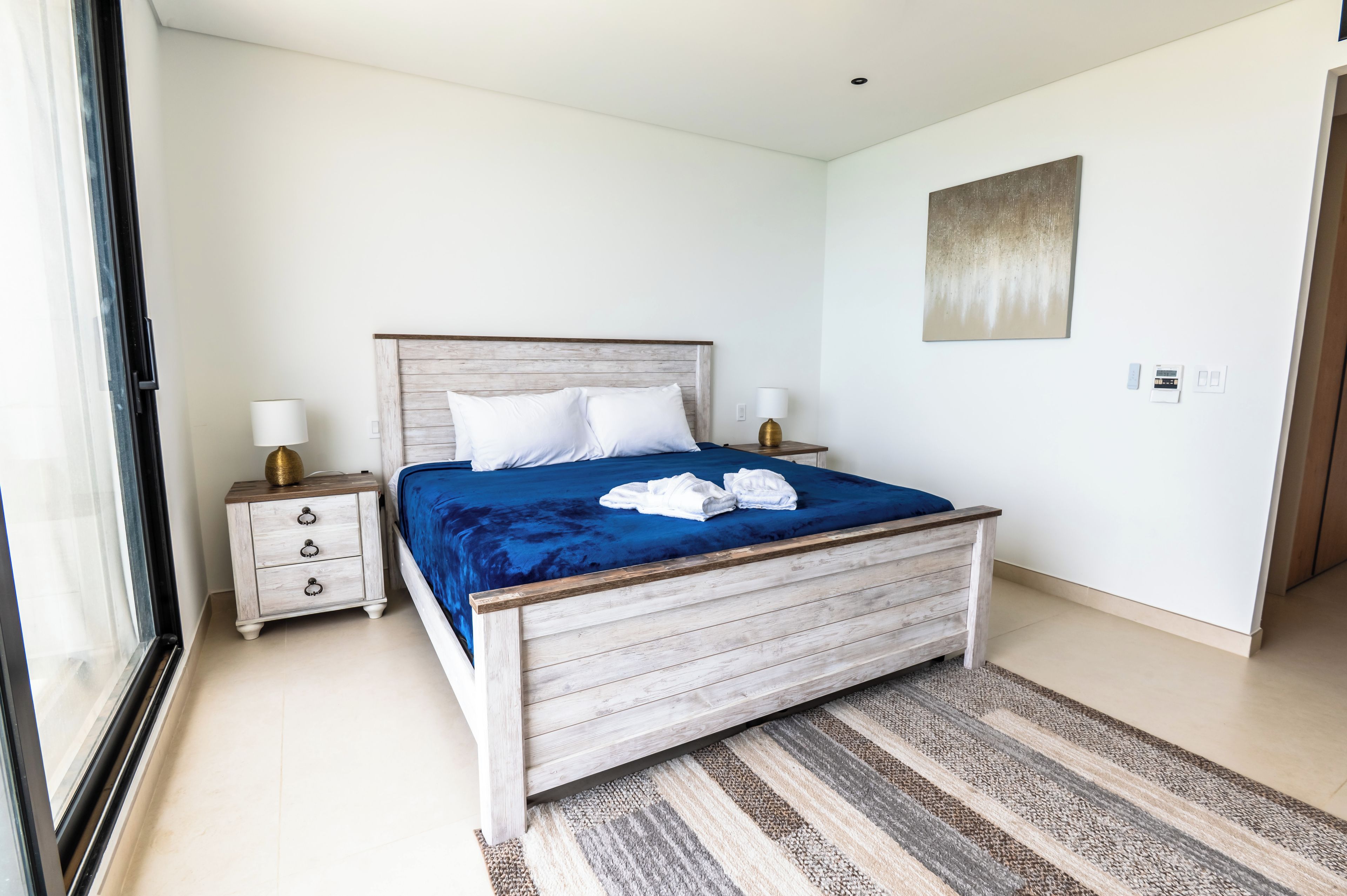 Un dormitorio con cama, mesitas de noche, lámpara, un cuadro y un termostato.