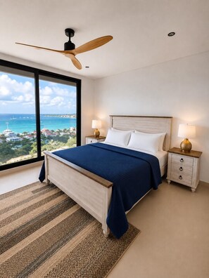 Ein Schlafzimmer mit großem Fenster, einem Bett, einem Nachttisch, einem Deckenventilator und Blick auf das Meer und Gebäude.