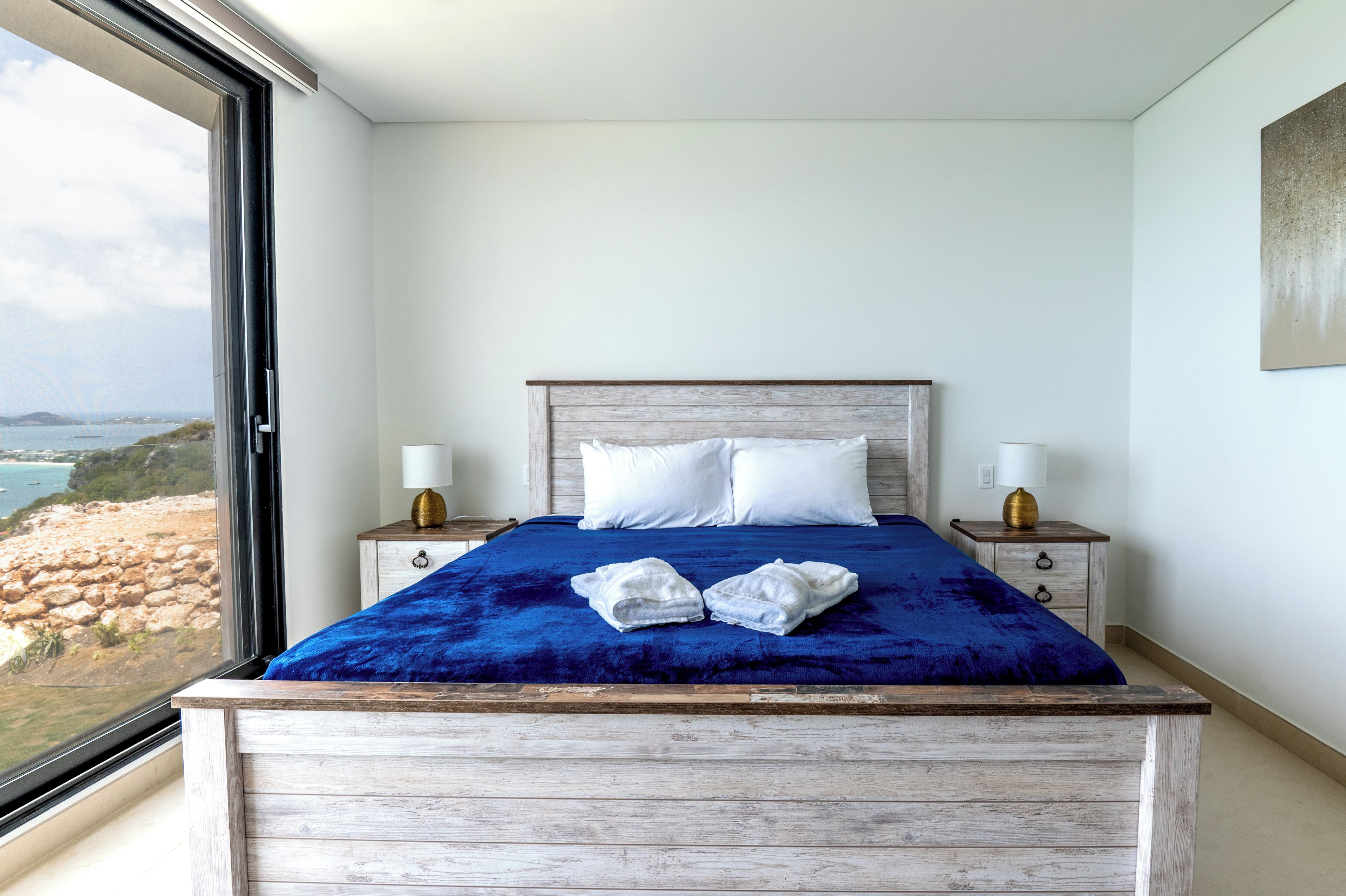 A bedroom with a large bed, two bedside tables, a view of the sea, and a wooden headboard.