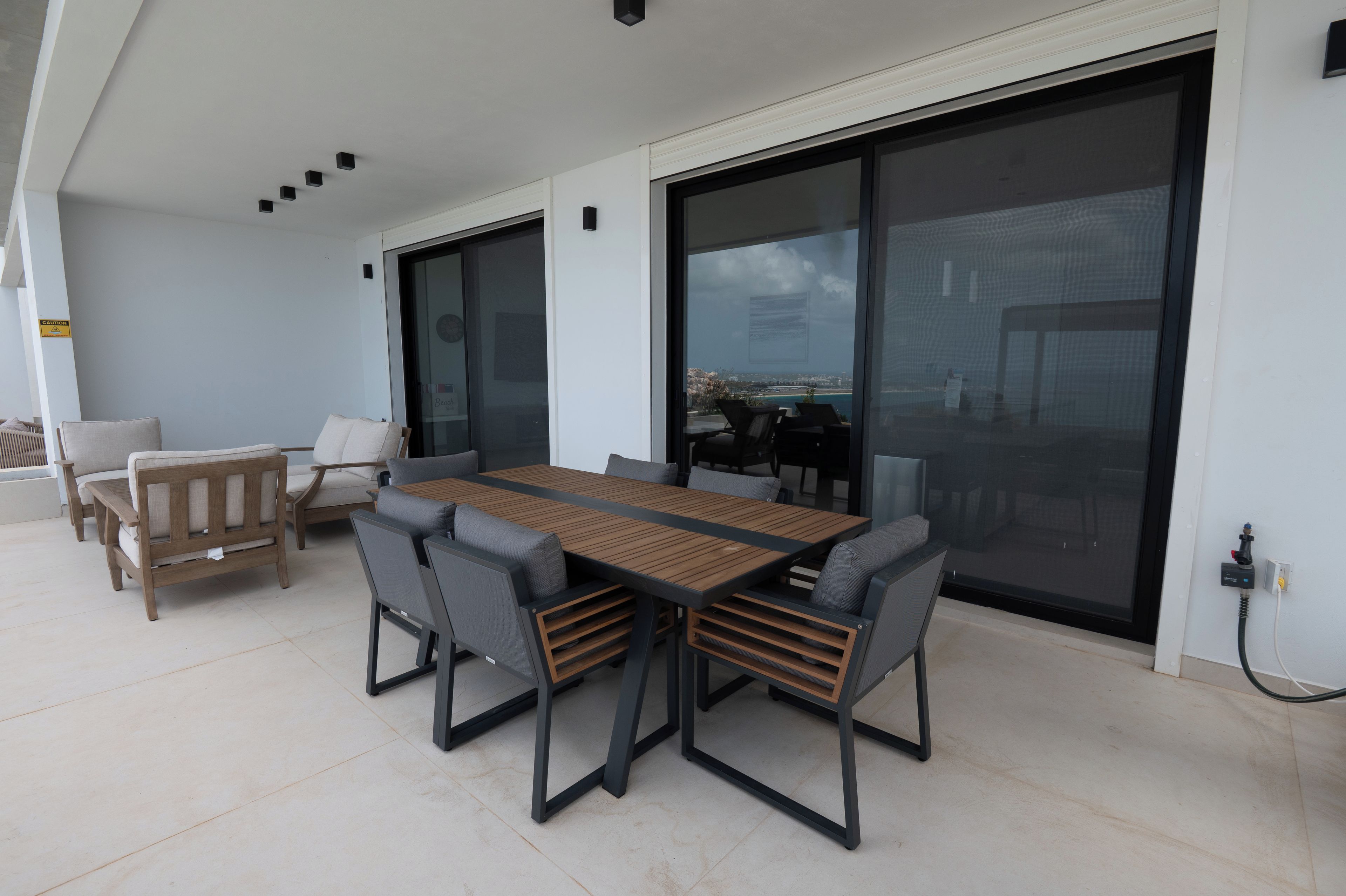An outdoor patio with a wooden table and chairs, a sofa, and a large sliding glass door leading to a view of the sea.