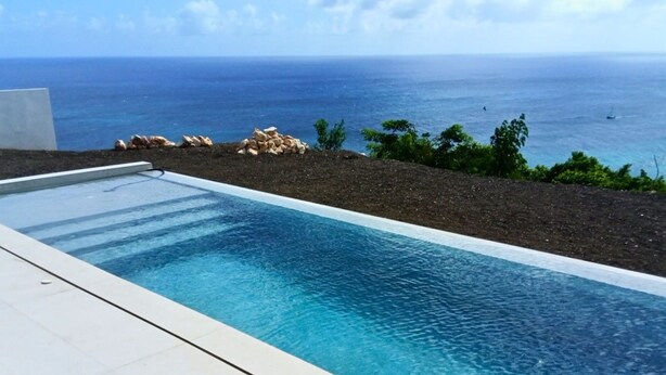 A rectangular pool with clear blue water, surrounded by a white tiled deck, with a view of the ocean and rocky terrain in the background. - Modern Pelican St. Maarten Escape with Pool, Views & Outdoor Lounge (Cole Bay)