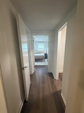 Ein schmaler Flur führt zu einem Schlafzimmer mit Bett, Nachttisch und einem Fenster mit Jalousien.
