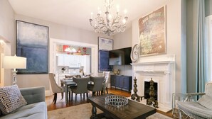 A living room with a chandelier, a fireplace, a dining area, and a kitchen in the background. - Spacious Historic 2BR w/ Courtyard, Parking, Walk to Forsyth Park (Savannah)