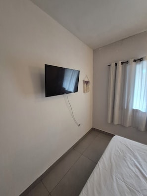 A room with a flat-screen TV mounted on the wall, a bed with white bedding, and a curtain on the window. - Cozy suite in Peruíbe, SP (Peruíbe)