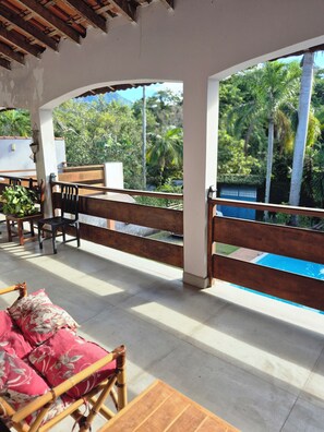 A covered outdoor seating area with wooden furniture and a view of a swimming pool. - Cozy suite in Peruíbe, SP (Peruíbe)