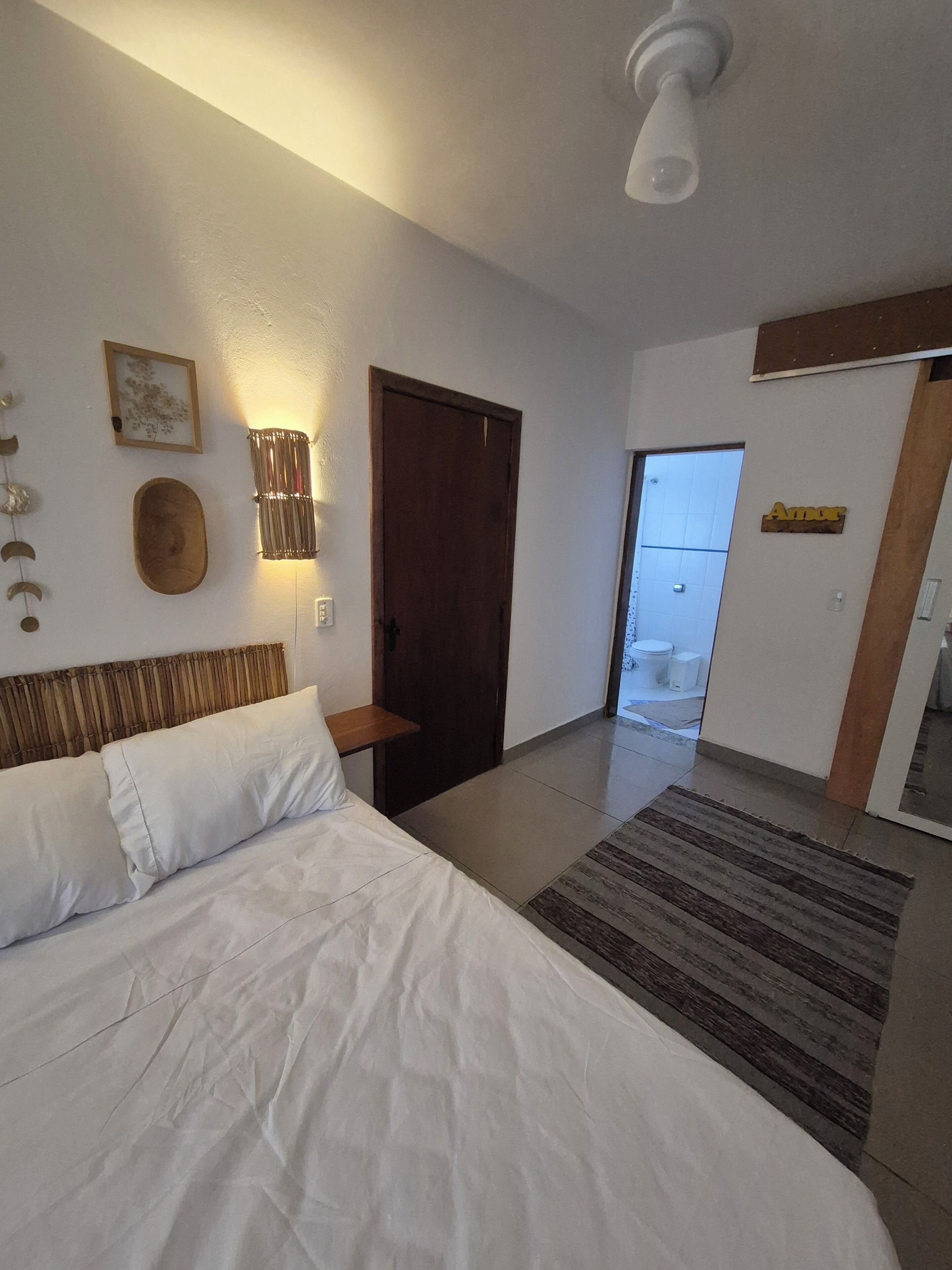 Une chambre à coucher avec un lit, des tables de chevet, une lampe fixée au mur, une porte en bois et une salle de bain visible à travers une porte ouverte.