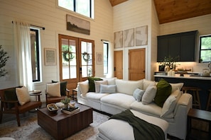 A cozy living room with a white sofa, a wooden coffee table, and a kitchen area with dark cabinets and a window. - Summit Sky: Charming Luxury Cabin with Treetop Views in Hochatown, Broken Bow (Broken Bow)