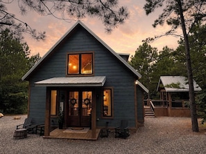 A small, single-story cabin with a porch, a visible interior with a fireplace, and a clear sky. - Summit Sky: Charming Luxury Cabin with Treetop Views in Hochatown, Broken Bow (Broken Bow)