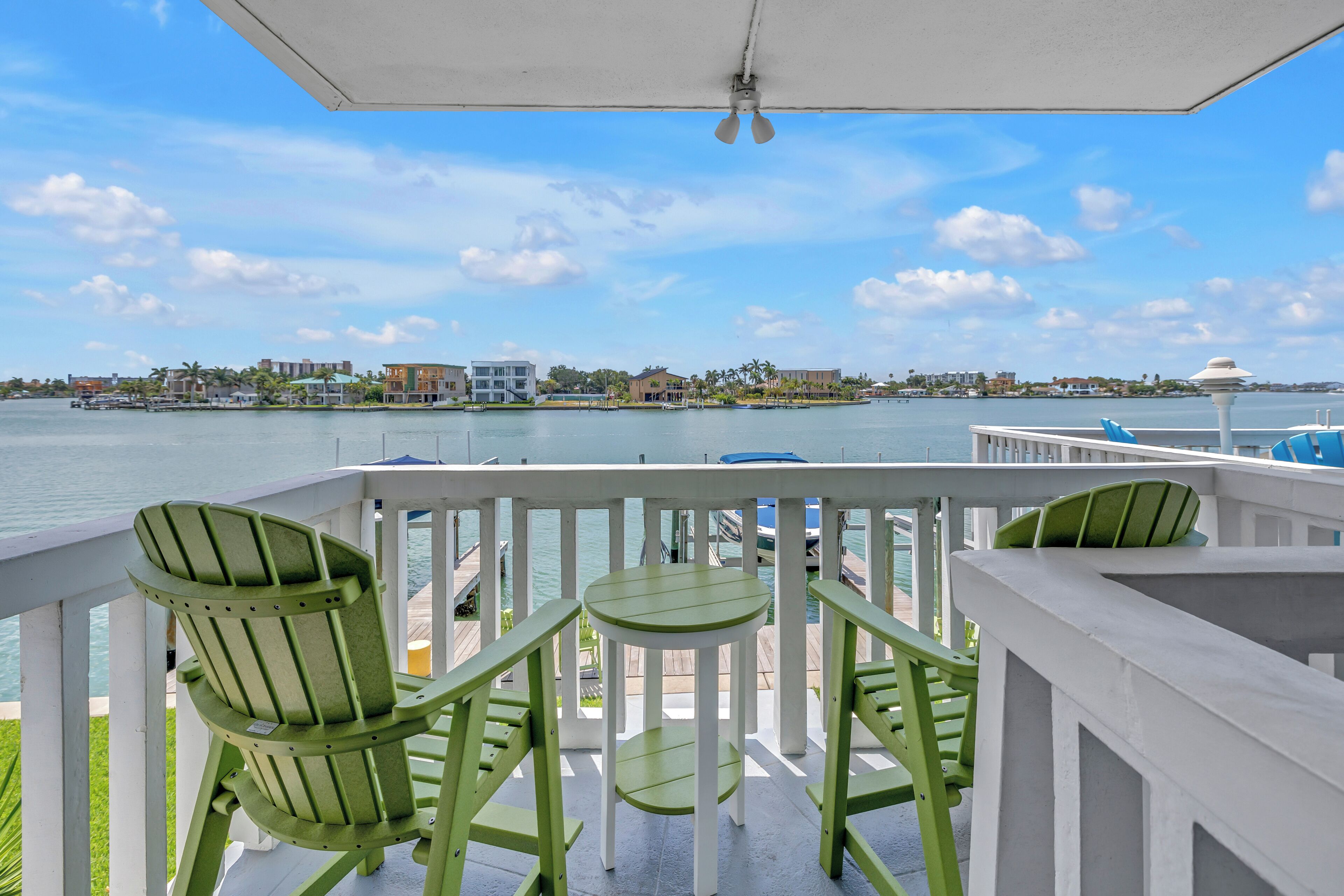 Un balcon agrémenté de fauteuils Adirondack verts et d’une petite table, donnant sur un front de mer avec des bâtiments et un ciel dégagé.