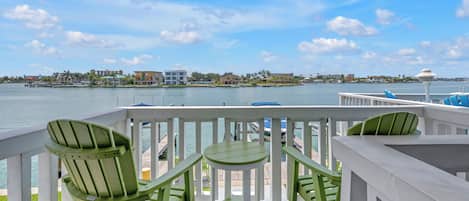A balcony with green Adirondack chairs and a small table, overlooking a waterfront with buildings and a clear sky.