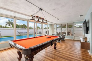 Ein Billardtisch in einem Raum mit großen Fenstern, einem Pool und einem Fernseher.