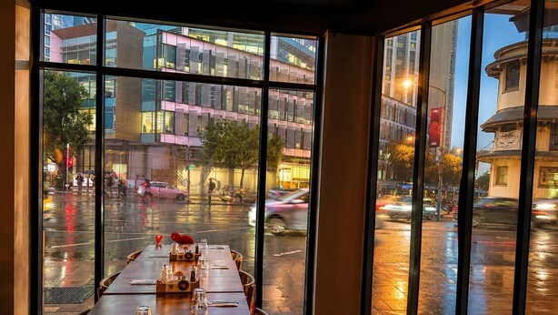 A restaurant with a long table set for dining, large windows offering a view of the city street. - Queen Street CBD Stay with Balcony and Amenities (Brisbane City)
