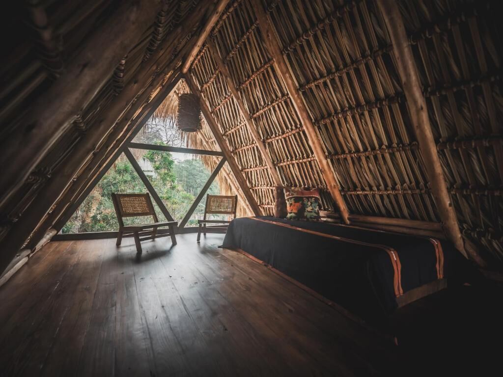A triangular wooden room with a thatched roof, wooden floor, and a bench.