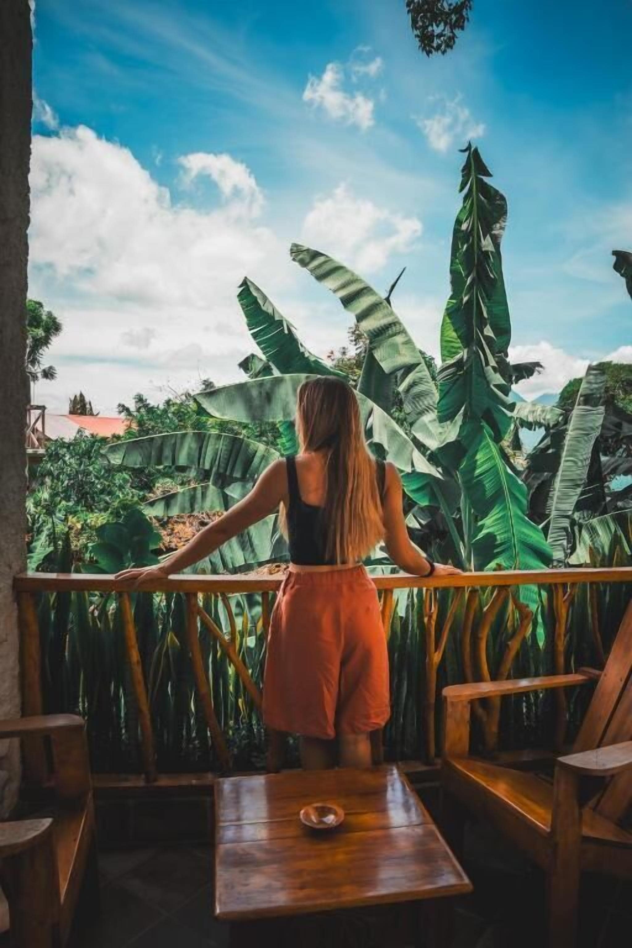 A person standing at a wooden table with a view of large banana leaves and a clear sky.