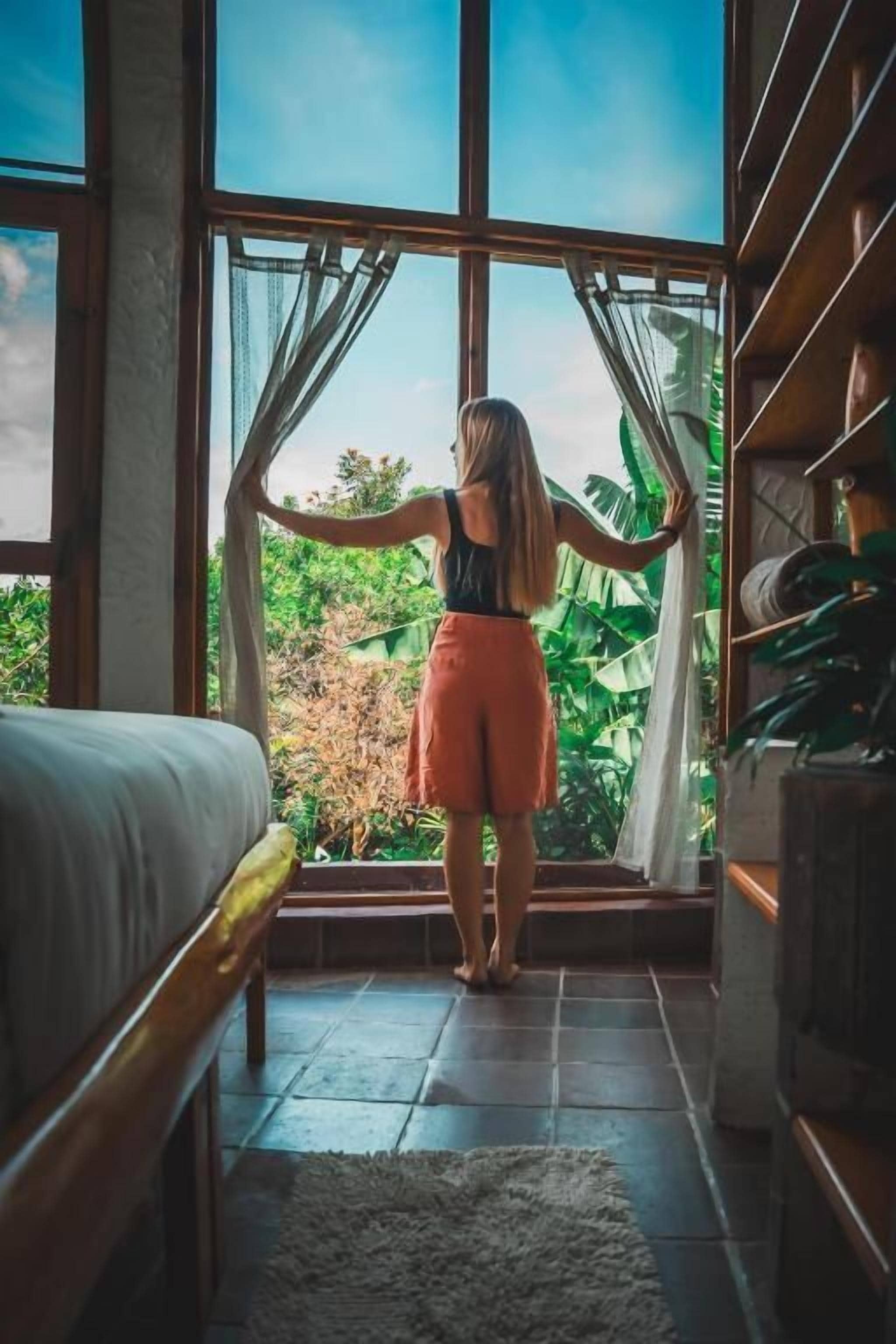 Una habitación con un ventanal grande, una cama y un estante de madera con plantas.
