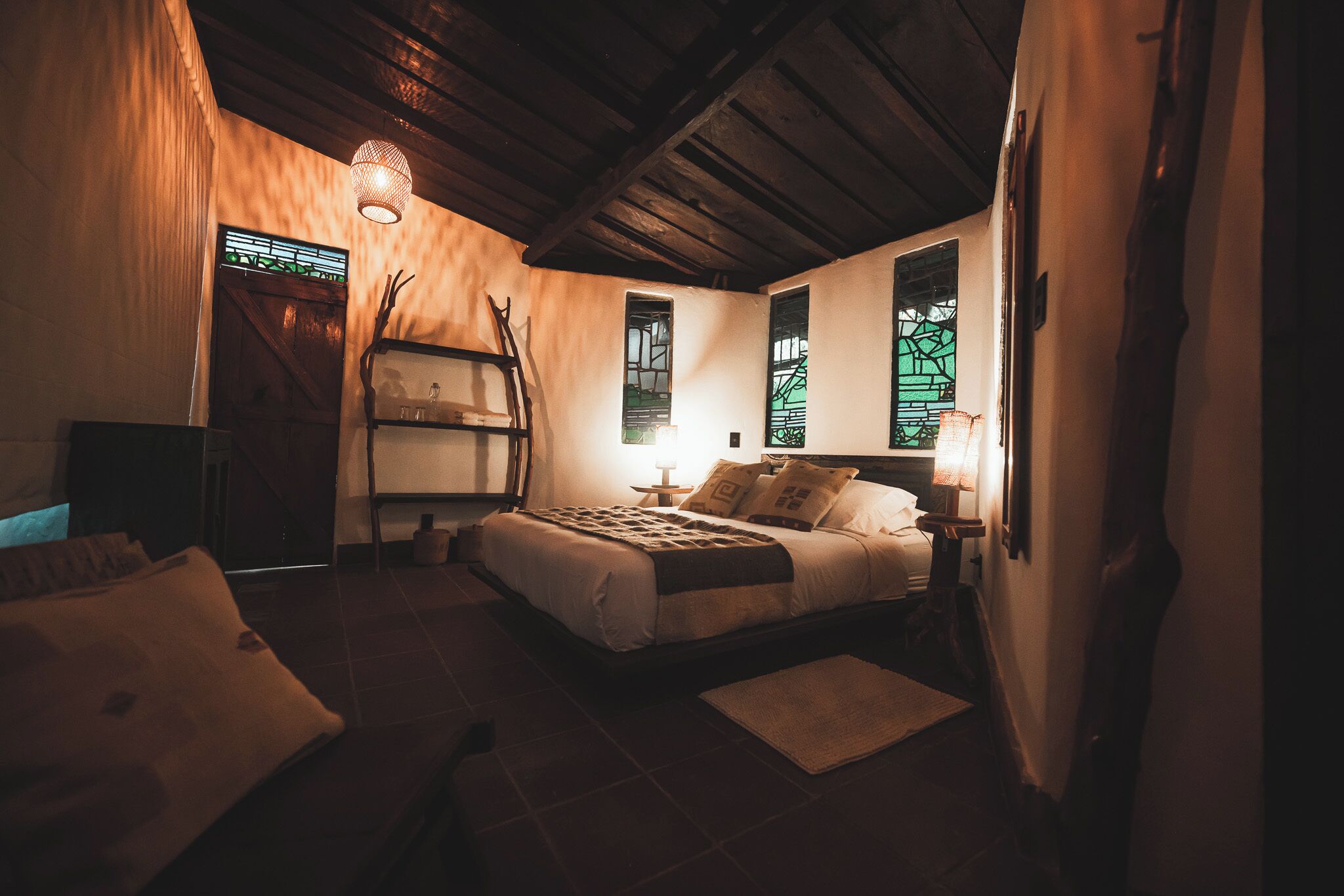 A dimly lit bedroom with a bed, a nightstand, a ladder, and a window with a view.
