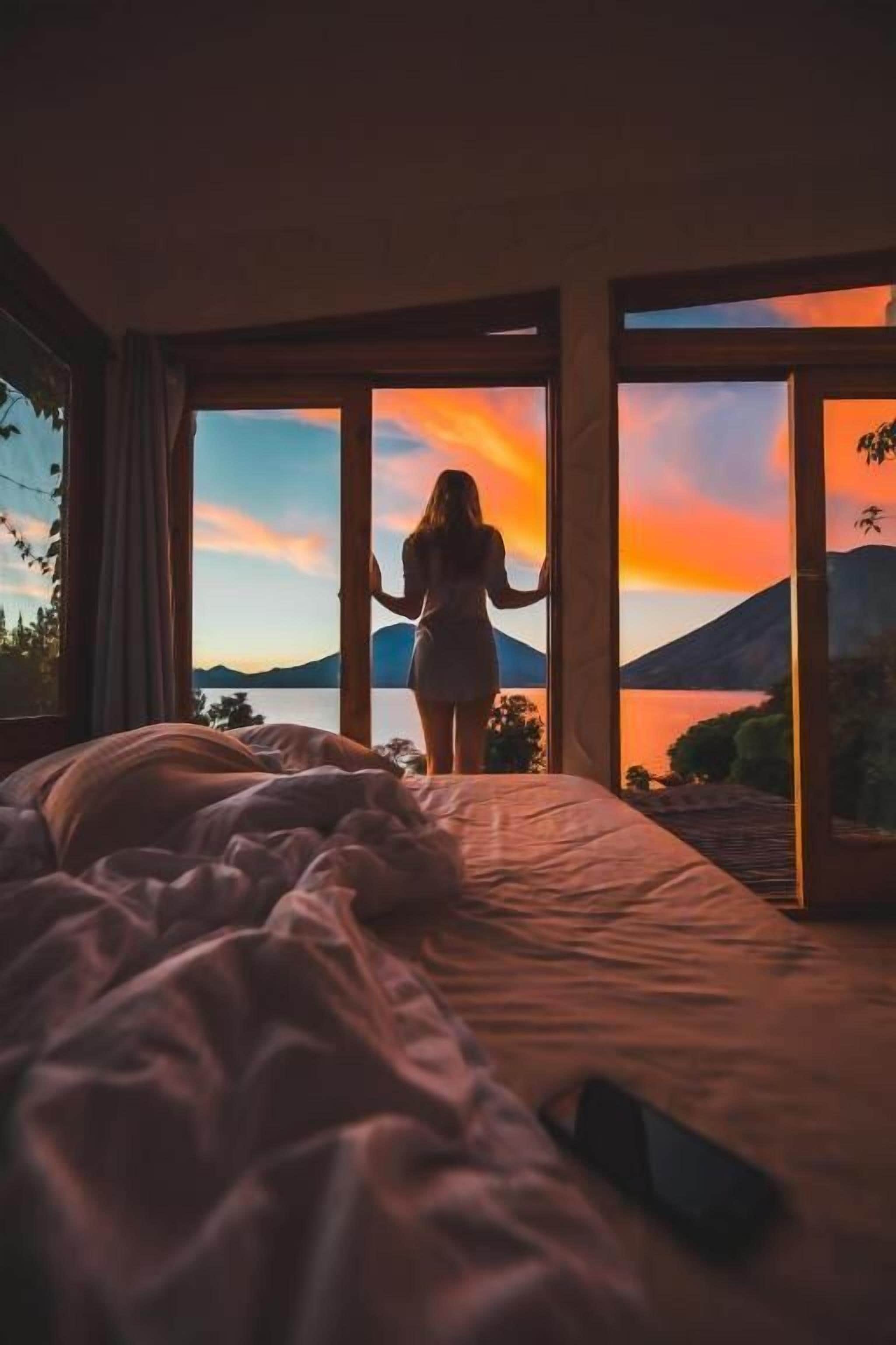 A room with a bed, a window, and a view of a sunset over a body of water.