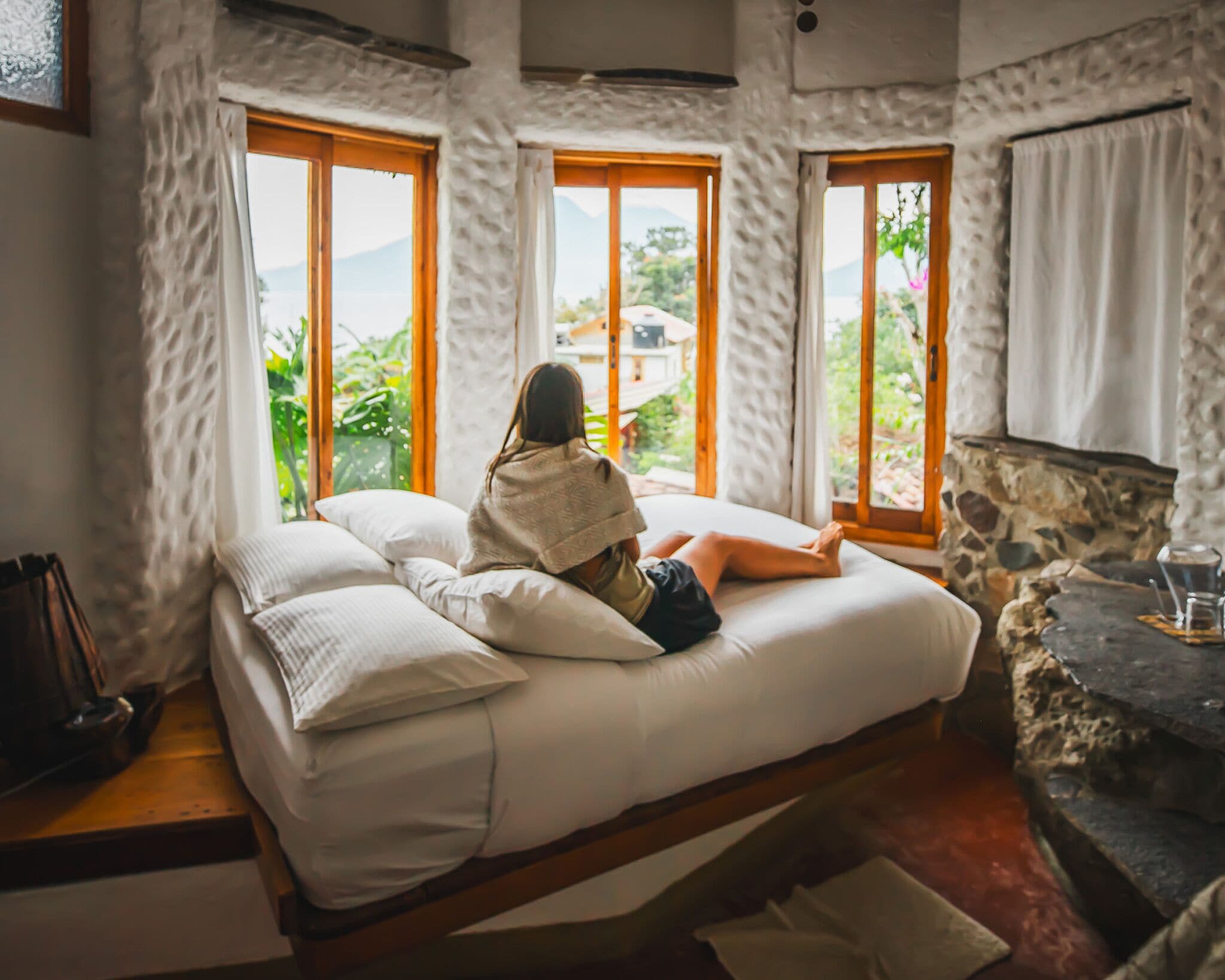 A room with a bed, large windows, a stone wall, and a table with a glass.