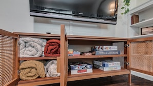 Une étagère en bois sur laquelle sont posés des couvertures pliées, des jeux de société et un téléviseur à écran plat fixé au mur.