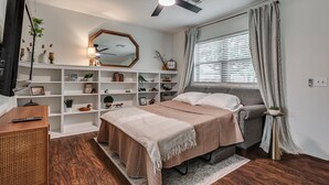Une chambre à coucher comprenant un lit, un bureau, une bibliothèque, un ventilateur de plafond et une fenêtre avec des rideaux.