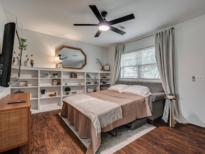A bedroom with a bed, a desk, a bookshelf, a ceiling fan, and a window with curtains.