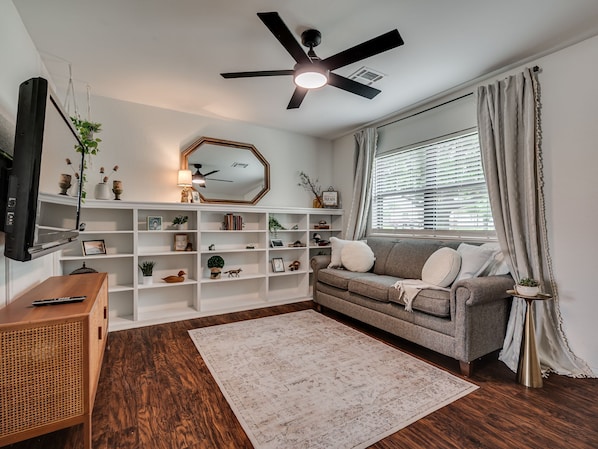 A living room with a sofa, a white bookshelf, a ceiling fan, and a window with curtains.