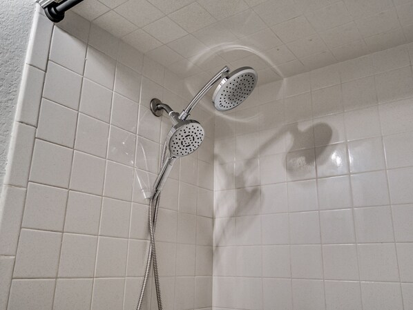 Une douche équipée d’un pommeau de douche à main et d’un pommeau fixé au mur, tous deux dotés d’une conception perforée, dans une pièce dont les murs sont recouverts de carreaux blancs.
