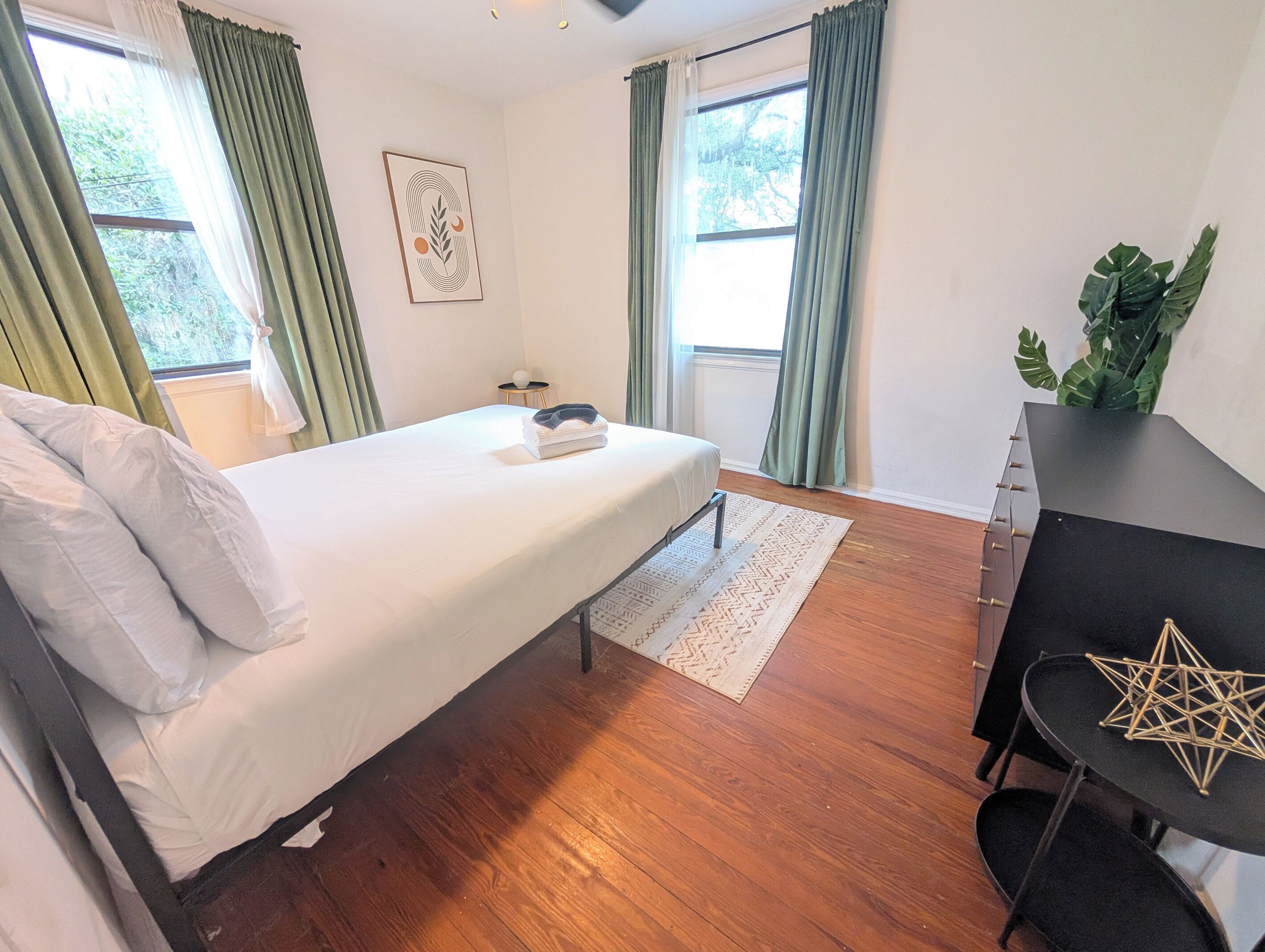 A bedroom with a bed, a desk, a chair, a plant, and a window with curtains.
