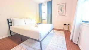 A bedroom with a bed, a nightstand, a wall-mounted lamp, a window with curtains, and a framed picture. - The Flagler Flat | San Marco Gem (Jacksonville)