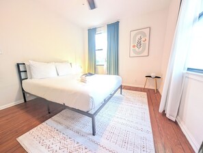 Ein Schlafzimmer mit einem Bett, einem Nachttisch, einer Wandlampe, einem Fenster mit Vorhängen und einem gerahmten Bild.