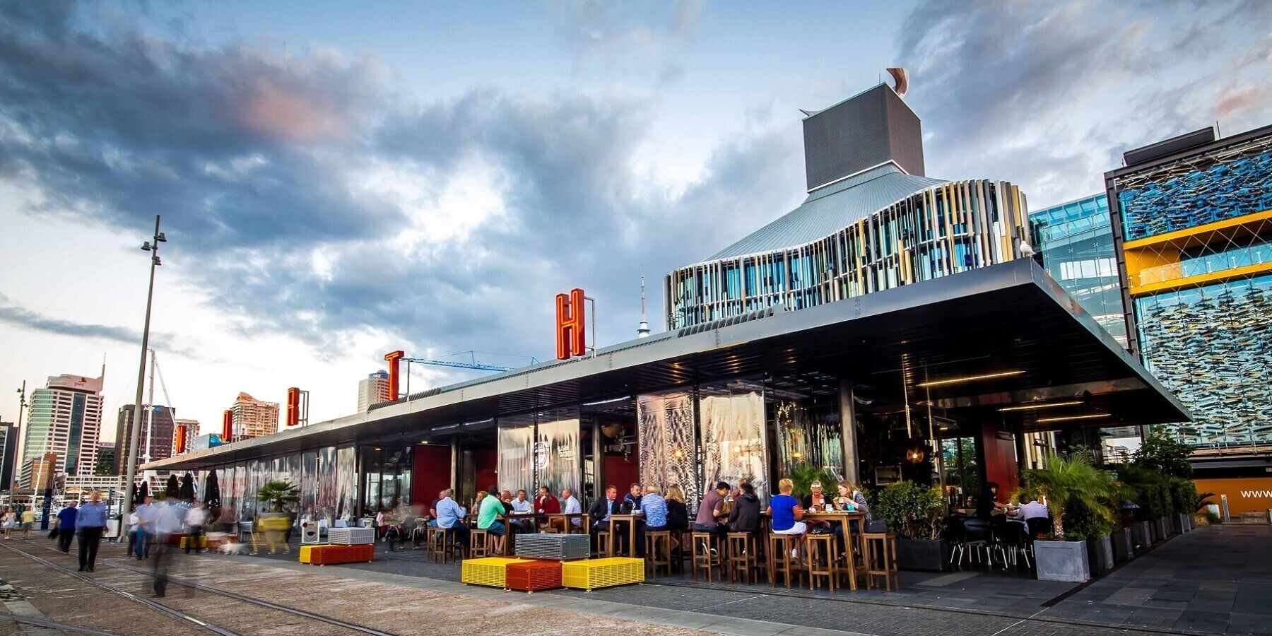 A modern building with a prominent 'H' sign, outdoor seating, and a view of the city skyline.