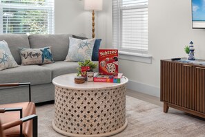 Ein Wohnzimmer mit grauem Sofa, einem runden Couchtisch, einem Bücherregal und einem Fenster mit Jalousien.