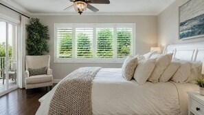 Ein Schlafzimmer mit Bett, einem Sessel, einem Fenster mit Jalousien, einem Deckenventilator und Blick ins Grüne.