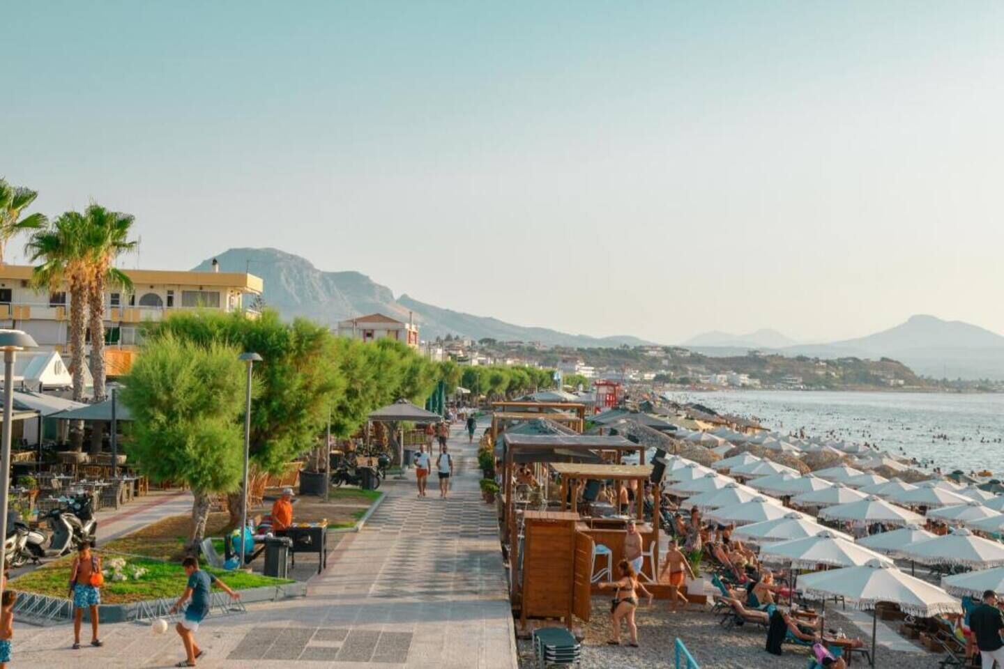 Un paseo marítimo con mesas al aire libre, palmeras y vistas al mar y a las montañas.