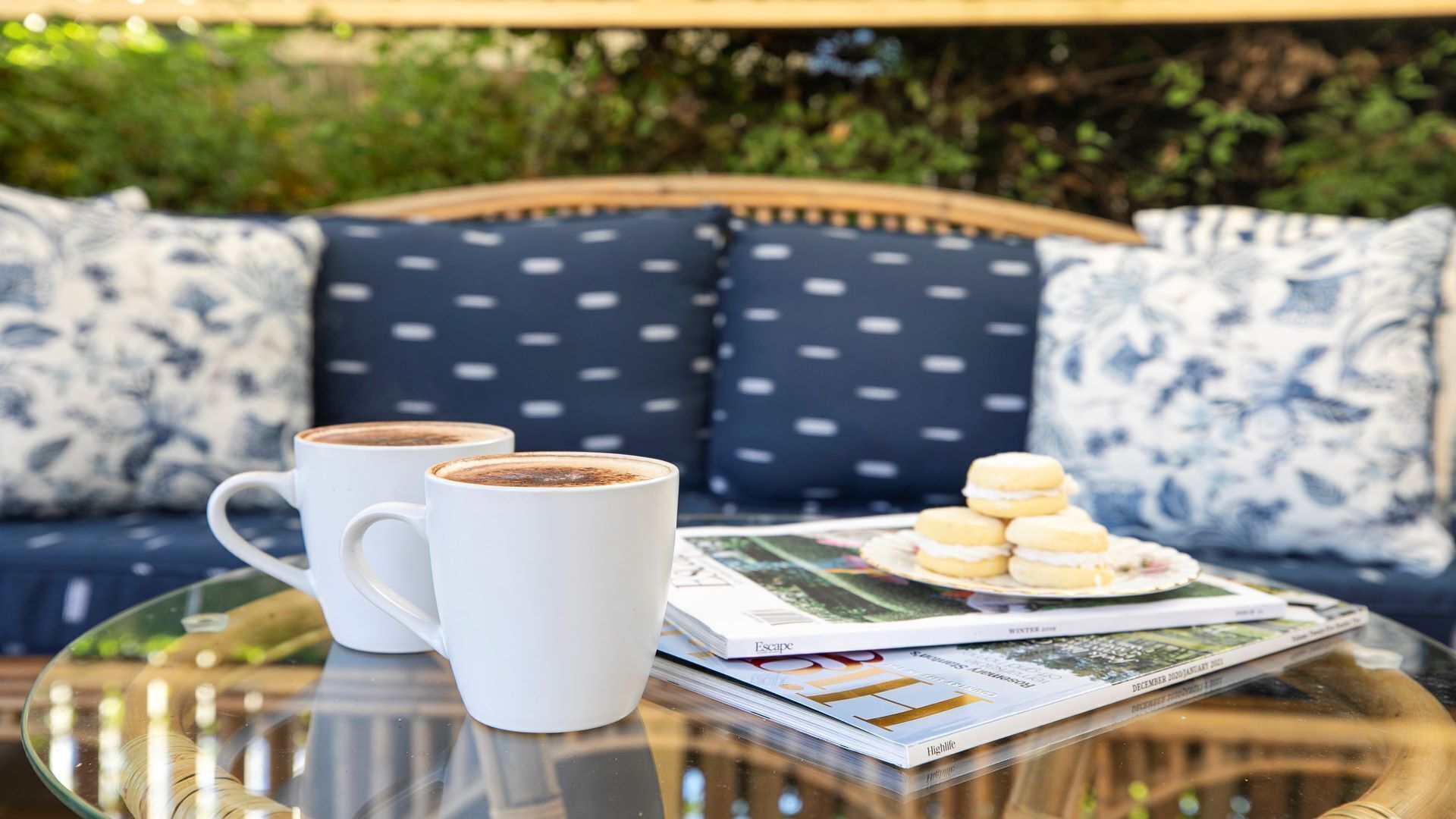 Una mesa de vidrio con dos tazas de café blancas, un plato de galletas y una revista.
