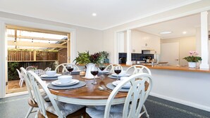A dining area with a round table set for six, white chairs, and a kitchen visible through an open doorway. - The Nook - central, cosy and well-equipped (Bowral)