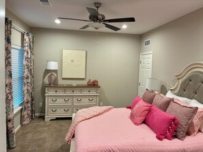 A bedroom with a bed, dresser, ceiling fan, and a window with curtains.