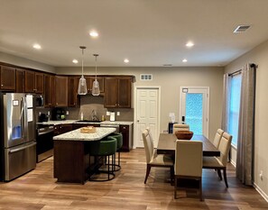 A modern kitchen with wooden cabinets, stainless steel appliances, a granite countertop island with bar stools, and a dining area with a table and chairs.
