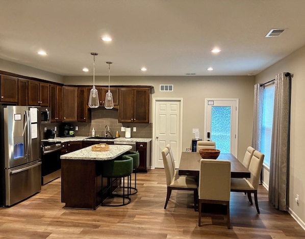 A modern kitchen with wooden cabinets, stainless steel appliances, a granite countertop island with bar stools, and a dining area with a table and chairs.