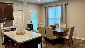 A kitchen with granite countertops, wooden cabinets, a dining area with a wooden table and chairs, and a window with curtains.