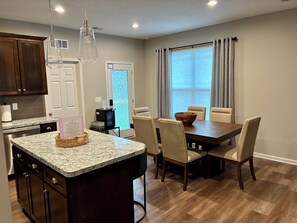 A kitchen with granite countertops, wooden cabinets, a dining area with a wooden table and chairs, and a window with curtains.