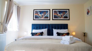 Una cama doble con cabecera azul, dos cuadros abstractos enmarcados, una lámpara de noche y una toalla enrollada sobre la cama.