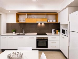 A modern kitchen with white cabinetry, a built-in oven, a microwave, and a wooden shelf above the sink. - 2BR Walk to James St | Balcony + Parking (Brisbane)