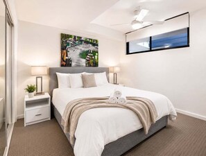 A modern hotel room with a bed, bedside tables, a ceiling fan, and artwork on the walls. - 2BR Walk to James St | Balcony + Parking (Brisbane)