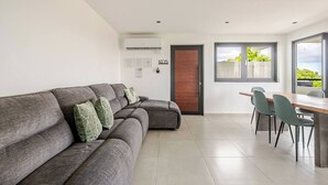 A modern living room with a grey sectional sofa, a dining area with a wooden table and chairs, and a large window offering a view of greenery. - Luxury Casa Kiarita • House on the Hill (San Juan)