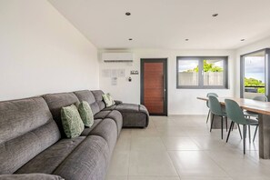A modern living room with a grey sectional sofa, a dining area with a wooden table and chairs, and a large window offering a view of greenery. - Luxury Casa Kiarita • House on the Hill (San Juan)