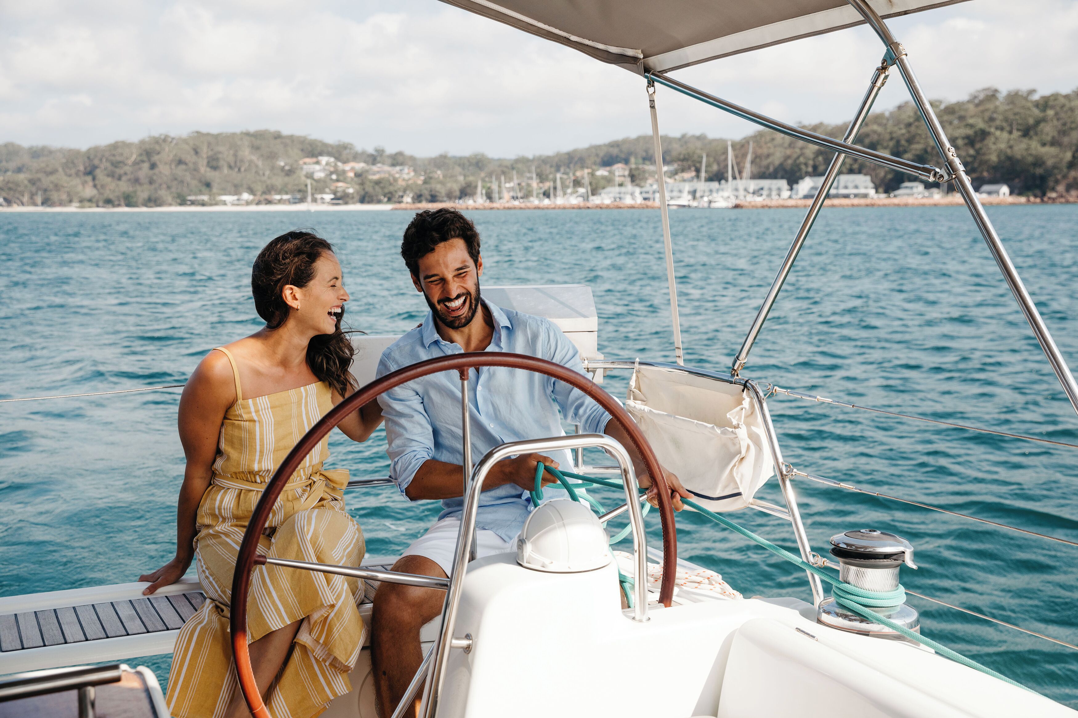 A couple on a sailboat with a view of a coastline and buildings in the background.